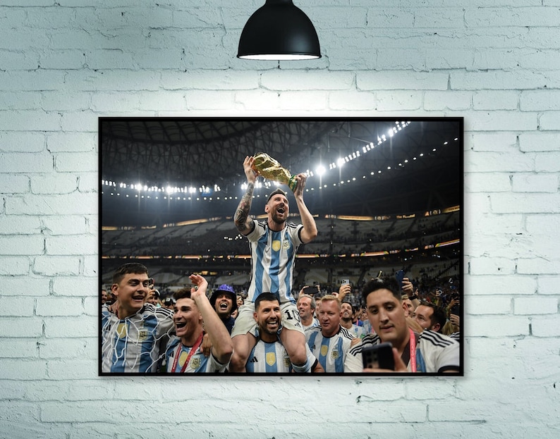 May include: A framed print of a soccer team celebrating a victory. The player in the center, wearing a blue and white striped jersey, holds a trophy aloft. Teammates surround him, some on shoulders, in a stadium setting.
