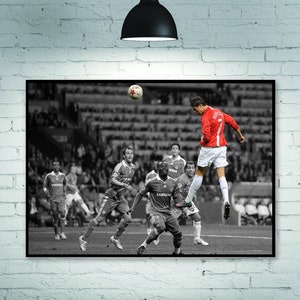 May include: A black and white photograph of a soccer player in a red jersey kicking a soccer ball in mid-air. The player is surrounded by other players in black and white jerseys. The background is a blurred image of a stadium.