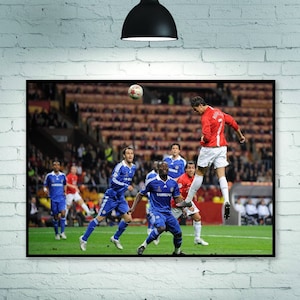 May include: A soccer player in a red jersey jumps in the air to head the ball.  Other players in blue jerseys are on the field.  The game is being played in a stadium with a large crowd.