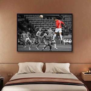 May include: A black and white photograph of a soccer player in a red jersey kicking a soccer ball. The player is in mid-air and the ball is in the air. The other players are in the background.