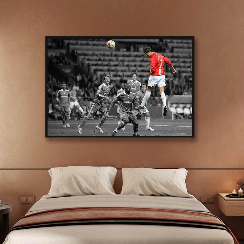 May include: A black and white photograph of a soccer player in a red jersey kicking a soccer ball. The player is in mid-air and the ball is in the air. The other players are in the background.