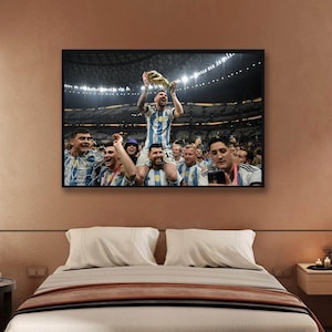 May include: Framed print of a soccer team celebrating a victory. The central figure, wearing a blue and white striped jersey, holds a trophy aloft. Teammates surround him, some on shoulders, in a stadium setting. The print is displayed above a bed.