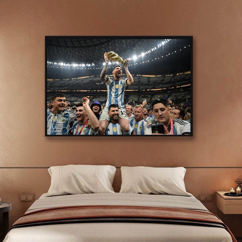 May include: Framed print of a soccer team celebrating a victory. The central figure, wearing a blue and white striped jersey, holds a trophy aloft. Teammates surround him, some on shoulders, in a stadium setting. The print is displayed above a bed.