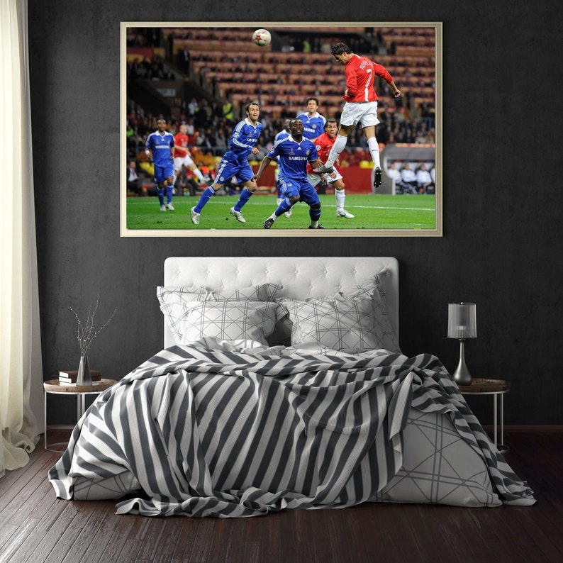 May include: A framed photograph of a soccer game with a player in a red jersey heading the ball. The player is surrounded by other players in blue jerseys. The photograph is hanging on a wall above a bed with a white headboard and striped bedding.