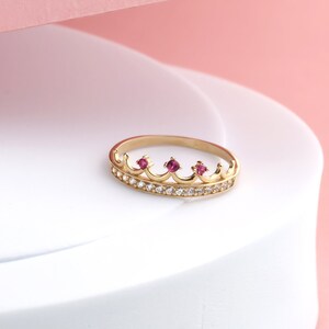 May include: A gold crown ring with three pink gemstones and a row of clear cubic zirconia stones.