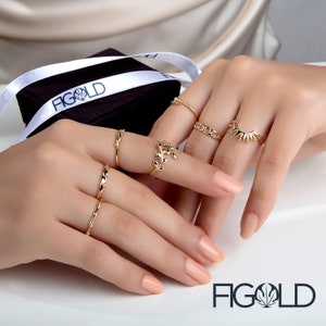 May include: A hand model wearing a variety of gold rings, including a ring with a leaf design, a ring with a twisted band, and a ring with a sunburst design. The rings are displayed on a black background with the word "FIGOLD" in white lettering.