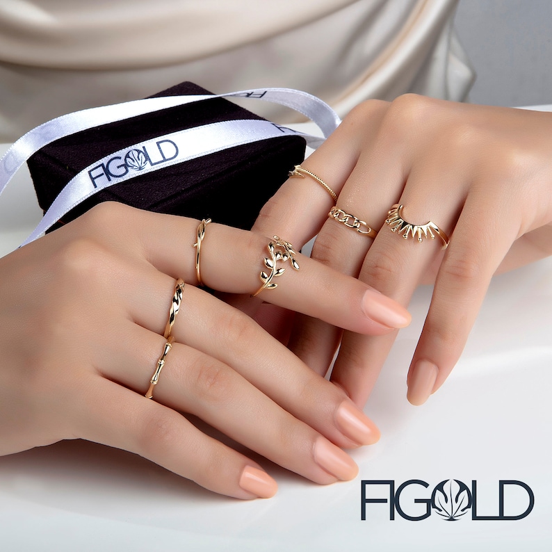 May include: A hand model wearing a variety of gold rings, including a ring with a leaf design, a ring with a twisted band, and a ring with a sunburst design. The rings are displayed on a black background with the word "FIGOLD" in white lettering.