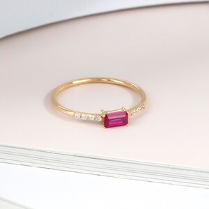 May include: A delicate gold ring featuring a rectangular, vibrant red gemstone as the focal point. The band is thin and adorned with small, sparkling white stones on one side, adding subtle elegance to this piece of jewelry.