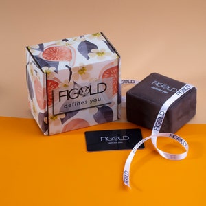 May include: A white box with a floral design and the text "FIGOLD defines you". The box is next to a brown velvet box with the same text on the side. A white ribbon with the same text is wrapped around the brown box. There is also a white gift card with the same text on a yellow surface.