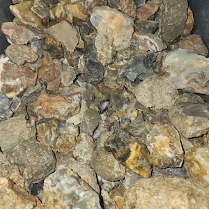May include: A collection of raw, uncut rocks in various sizes and shapes. The stones display a range of colors, including beige, brown, gray, and yellow. Some rocks have a polished appearance, while others are rough and textured.