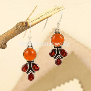 May include: A pair of silver dangle earrings with orange and red gemstones. The earrings feature a round orange gemstone at the top and four teardrop-shaped red gemstones hanging below.