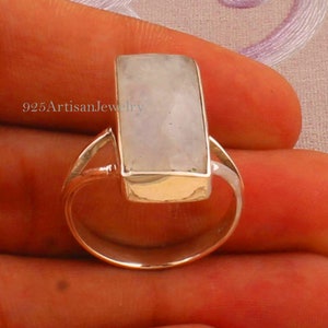 Puede incluir: Un anillo de plata con una piedra preciosa rectangular blanca. El anillo está engastado en una banda simple con un borde ligeramente elevado alrededor de la piedra preciosa.