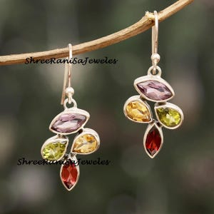 Puede incluir: Pendientes colgantes de plata con un grupo de piedras preciosas de colores. Cada pendiente presenta una mezcla de piedras en forma de lágrima y ovaladas de color púrpura, amarillo, verde y rojo. Los pendientes están suspendidos de un gancho de plata.