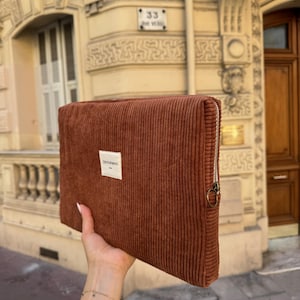 May include: A rectangular, rust-colored corduroy clutch with a silver zipper and a small white label that reads "TENTOPMENT". The clutch is held up in front of a building with a light-colored facade.