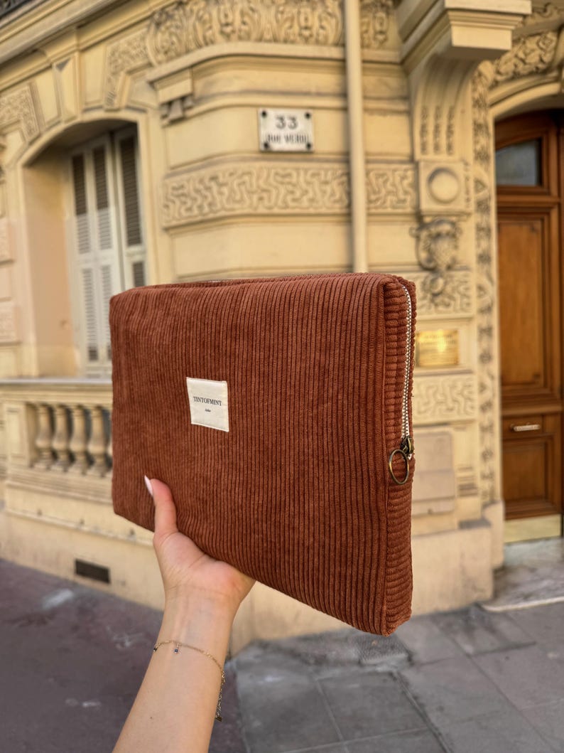 May include: A rectangular, rust-colored corduroy clutch with a silver zipper and a small white label that reads "TENTOPMENT". The clutch is held up in front of a building with a light-colored facade.