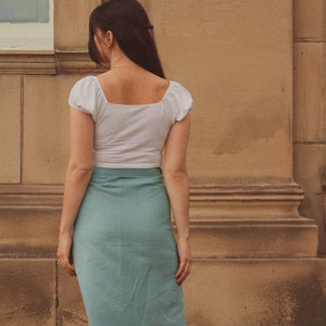 Pu&ograve; includere: Una donna indossa un top bianco corto con scollo quadrato e una gonna blu chiaro. La gonna ha uno stile a portafoglio ed &egrave; aderente.