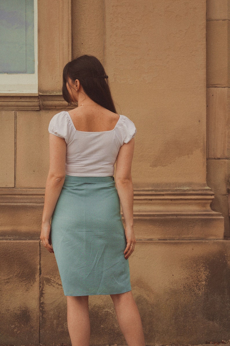 Pu&ograve; includere: Una donna indossa un top bianco corto con scollo quadrato e una gonna blu chiaro. La gonna ha uno stile a portafoglio ed &egrave; aderente.