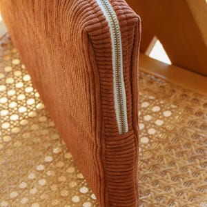 May include: A rectangular, rust-colored corduroy pouch with a white zipper. The pouch is resting on a woven rattan chair. The zipper is a light gold color.