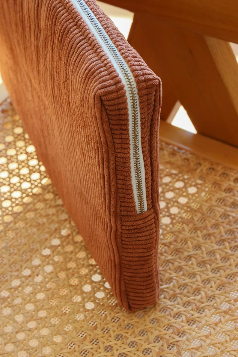 May include: A rectangular, rust-colored corduroy pouch with a white zipper. The pouch is resting on a woven rattan chair. The zipper is a light gold color.