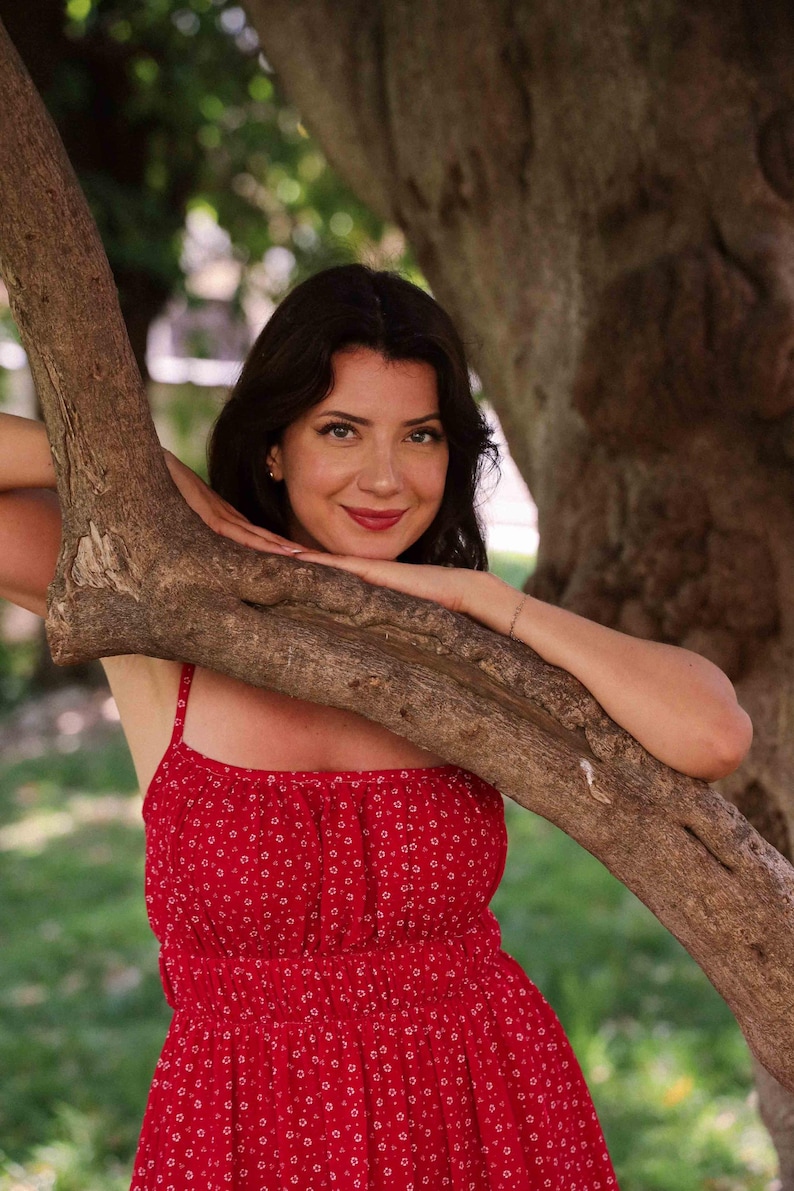 May include: A bright red sleeveless dress featuring a small white floral pattern, a gathered square neckline, and a smocked bodice. The garment has thin shoulder straps and a flared skirt, providing a simple and consistent design throughout the piece.