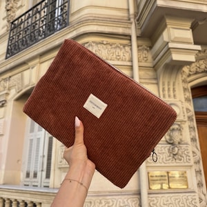 May include: A rectangular, rust-colored corduroy clutch with a zipper closure. A small white label with the text "TINTOFMINY" is affixed to the front. The clutch is held in front of a building with ornate architectural details.