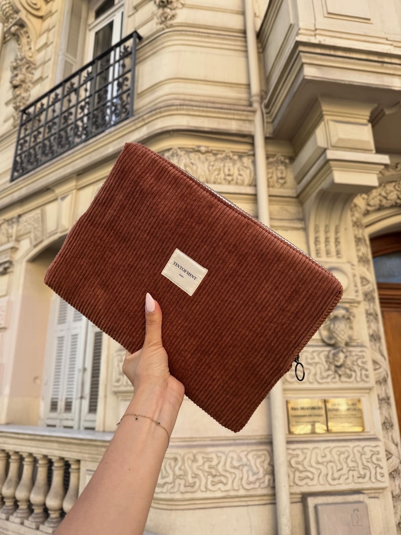 May include: A rectangular, rust-colored corduroy clutch with a zipper closure. A small white label with the text "TINTOFMINY" is affixed to the front. The clutch is held in front of a building with ornate architectural details.