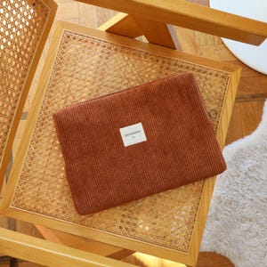 May include: A rust-colored corduroy laptop sleeve with a rectangular label that reads "TINTORMENT" is placed on a woven rattan chair. The chair has a light wood frame. The background includes a wooden floor and a white table.