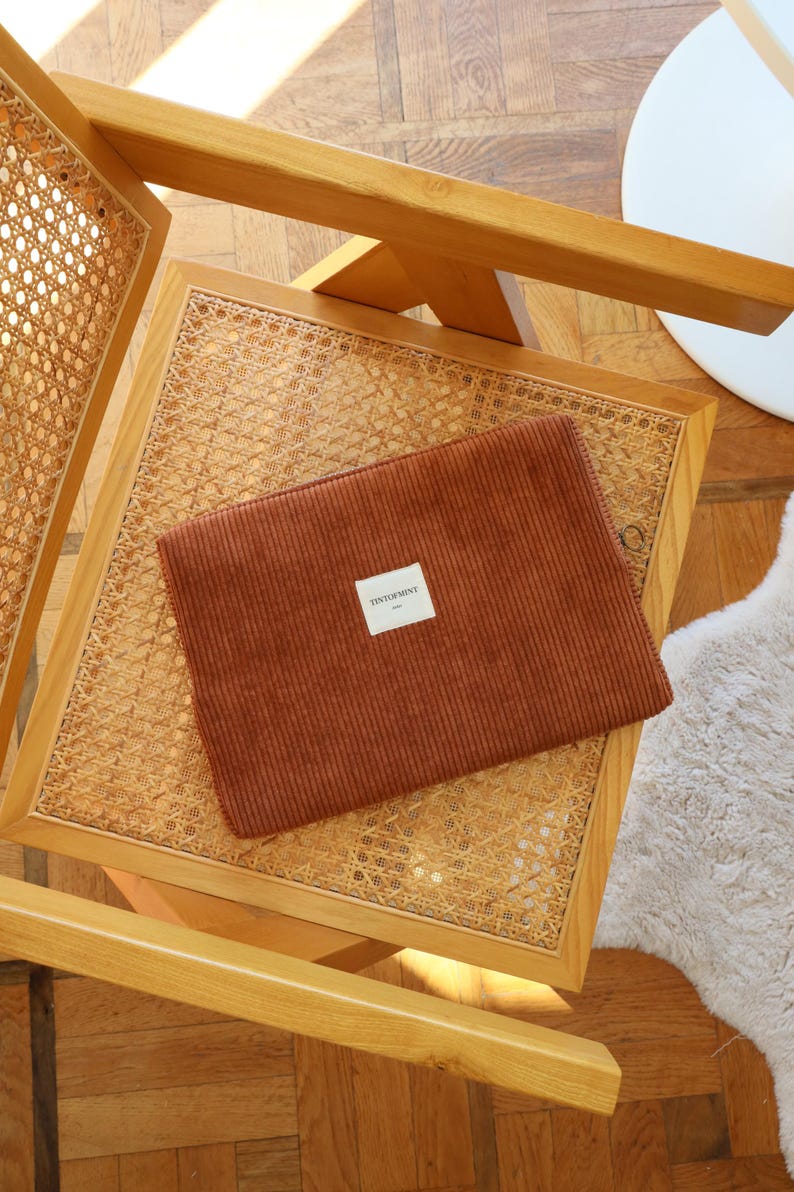 May include: A rust-colored corduroy laptop sleeve with a rectangular label that reads "TINTORMENT" is placed on a woven rattan chair. The chair has a light wood frame. The background includes a wooden floor and a white table.