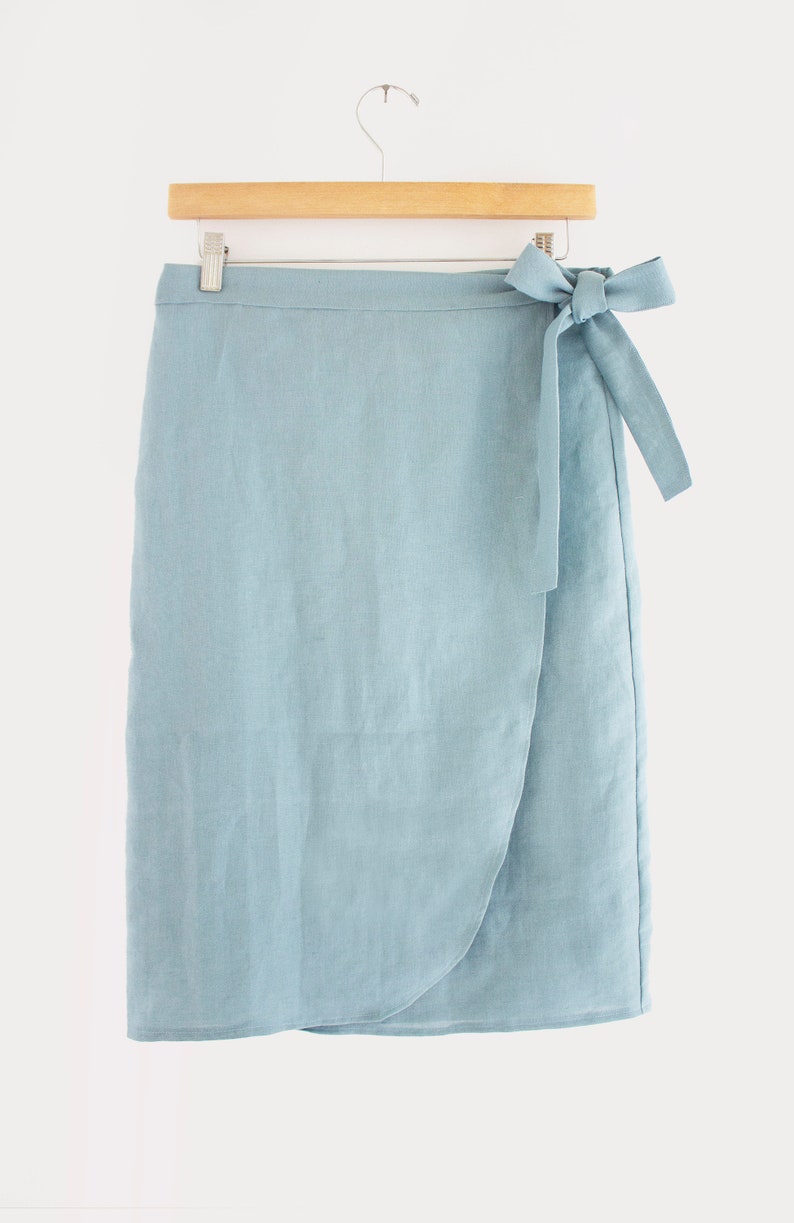 Pu&ograve; includere: Una gonna a portafoglio in lino blu chiaro con una cintura da annodare. La gonna &egrave; lunga fino al ginocchio e ha un design semplice ed elegante.
