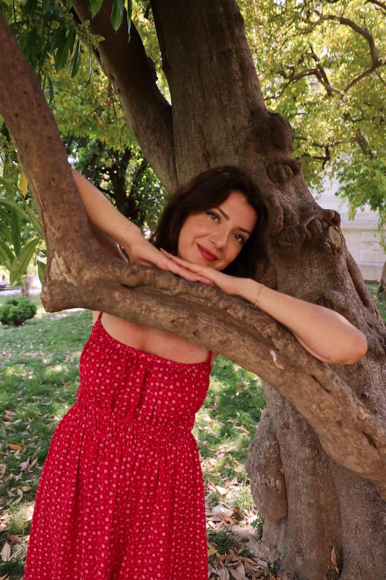 May include: A bright red sleeveless dress featuring a small white floral pattern, a square neckline, and a gathered, elasticised bodice that transitions into a flowing skirt. The garment is constructed with thin shoulder straps and a simple silhouette.