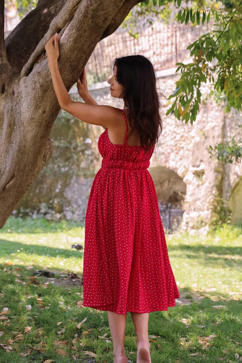 May include: A vibrant red midi dress featuring a delicate white floral pattern, thin shoulder straps, a fitted bodice with a gathered waistline, and a flowing, pleated skirt that falls below the knees, showcasing a simple and classic silhouette.