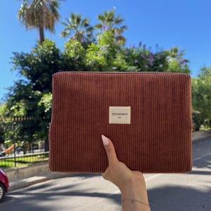 May include: A brown corduroy clutch with a rectangular shape is held up against a backdrop of palm trees and a bright blue sky. The clutch has a small white label that reads "TENTOFMINT".