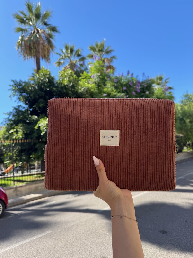 May include: A brown corduroy clutch with a rectangular shape is held up against a backdrop of palm trees and a bright blue sky. The clutch has a small white label that reads "TENTOFMINT".