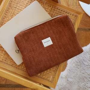 May include: A brown corduroy laptop sleeve with a silver zipper and a small white label that reads "TINTOFMINT". The sleeve is resting on a wooden chair with a woven seat, next to a laptop.