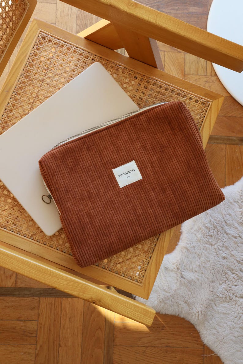 May include: A brown corduroy laptop sleeve with a silver zipper and a small white label that reads "TINTOFMINT". The sleeve is resting on a wooden chair with a woven seat, next to a laptop.