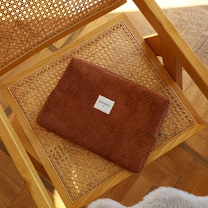 May include: A brown corduroy laptop sleeve with a small white label that reads "TINTORMINT" is placed on a wooden chair with a woven cane seat. The chair is made of light-colored wood.