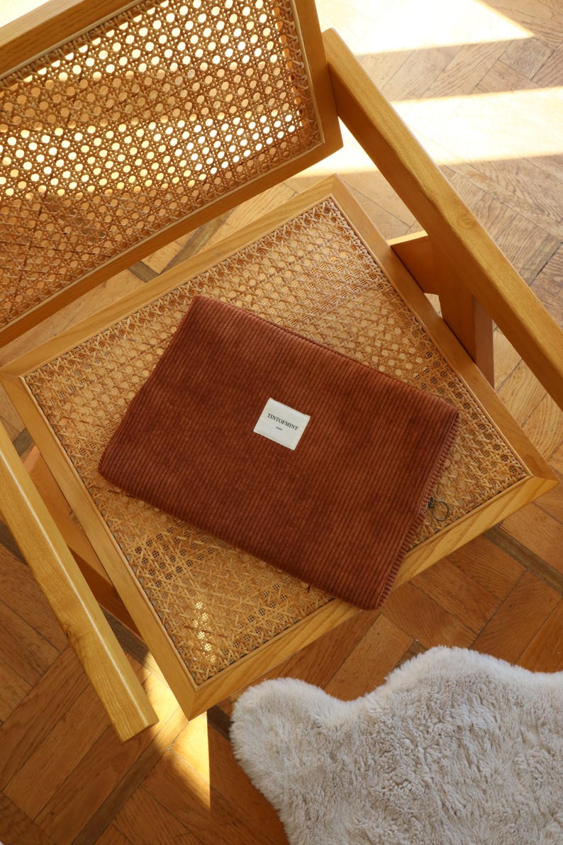 May include: A brown corduroy laptop sleeve with a small white label that reads "TINTORMINT" is placed on a wooden chair with a woven cane seat. The chair is made of light-colored wood.