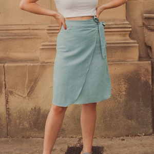 Pu&ograve; includere: Una donna indossa un top bianco corto con maniche a sbuffo e una gonna avvolgente blu chiaro. Indossa sandali con tacco blu chiaro.