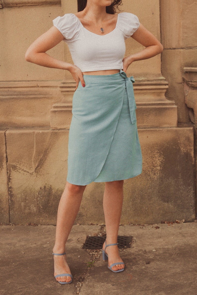 Pu&ograve; includere: Una donna indossa un top bianco corto con maniche a sbuffo e una gonna avvolgente blu chiaro. Indossa sandali con tacco blu chiaro.