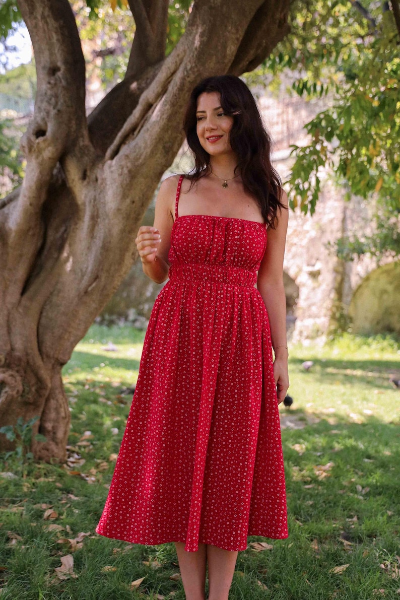 May include: A midi-length red dress featuring a small white floral pattern, a square neckline, thin shoulder straps, and a gathered elasticated waistline that flows into a full skirt. The garment is constructed with a fitted bodice and a flared hem.