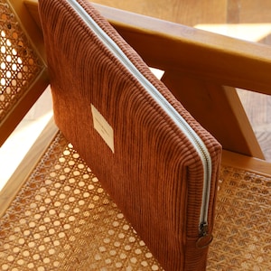 May include: A rust-colored corduroy laptop sleeve with a white zipper and a small rectangular label. The sleeve is rectangular and sits on a wooden chair with a woven seat.