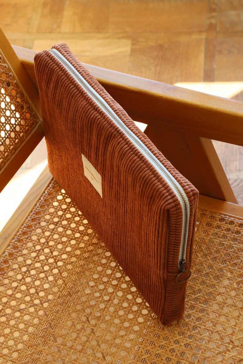 May include: A rust-colored corduroy laptop sleeve with a white zipper and a small rectangular label. The sleeve is rectangular and sits on a wooden chair with a woven seat.