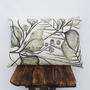 May include: Rectangular decorative pillow with a botanical design. The pillow features a cream-colored background with a pattern of olive green and charcoal-colored leaves and stems. The pillow is resting on a small, dark wooden stool.