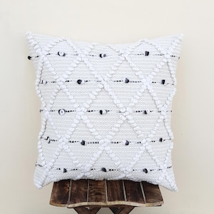 Peut inclure: Un coussin carré blanc cassé avec un motif en losanges texturé. Le coussin présente de petits accents de couleur foncée et est présenté sur un petit tabouret en bois. Le design du coussin ajoute une touche de texture et d'intérêt visuel.