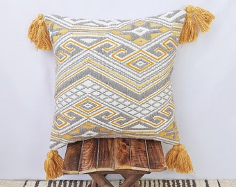 May include: Square decorative pillow with a geometric pattern in gray, white, and yellow. The pillow has yellow tassels on each corner. The pillow is placed on a small wooden stool.