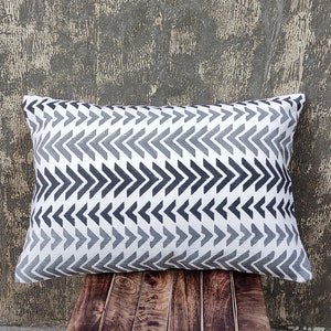 May include: Rectangular decorative pillow with a white background and a repeating chevron pattern in shades of gray and navy blue. The pillow is placed on a wooden surface.