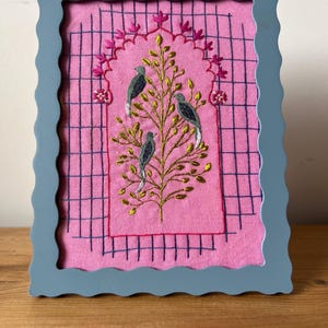 May include: A framed embroidery featuring a pink background with a grid pattern. A gold tree with three birds is embroidered in the center. The frame is a light blue color with a scalloped edge.