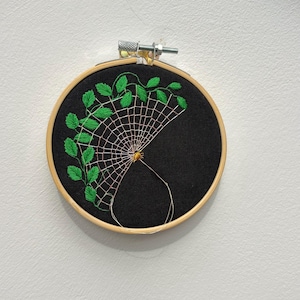 May include: A circular wooden embroidery hoop containing black fabric with a hand-stitched design. The embroidery features a white spider web segment attached to a vine with bright green leaves and a small golden spider at the centre point.