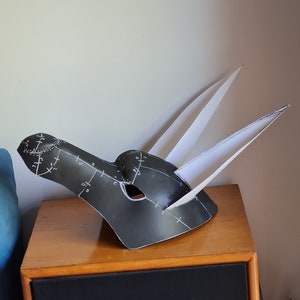 May include: A black paper mache mask with white markings and two white paper ears. The mask is sitting on a wooden table.