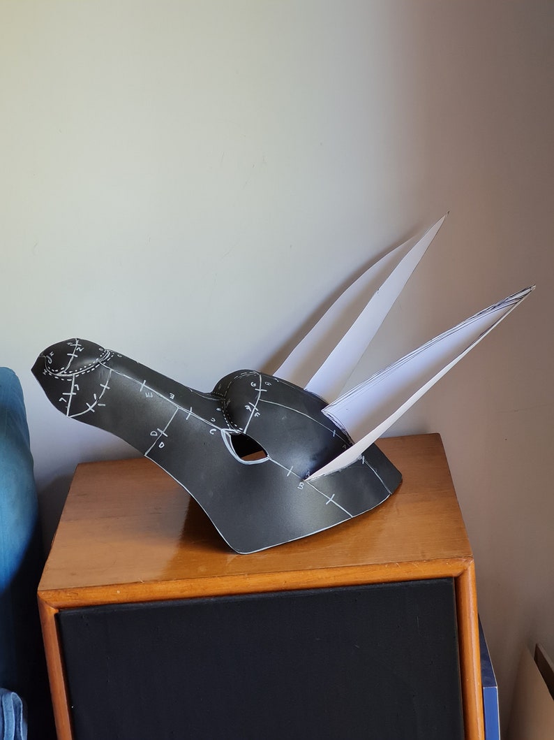 May include: A black paper mache mask with white markings and two white paper ears. The mask is sitting on a wooden table.
